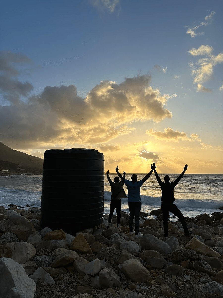 Three people with arms raised beside a Makulu tank on the rocks at sunset